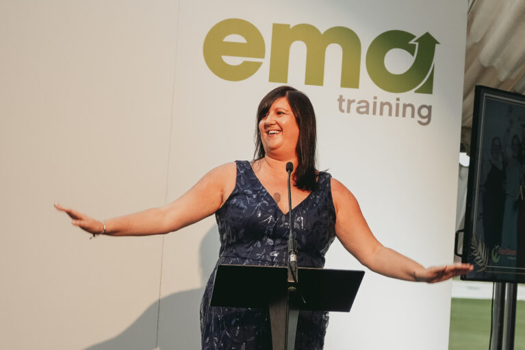 Managing Director, Tracey Mosley, on stage at the EMA Awards 2023. Tracey is wearing a navy blue sparkly dress and is standing infront of a white background with the green ema logo.